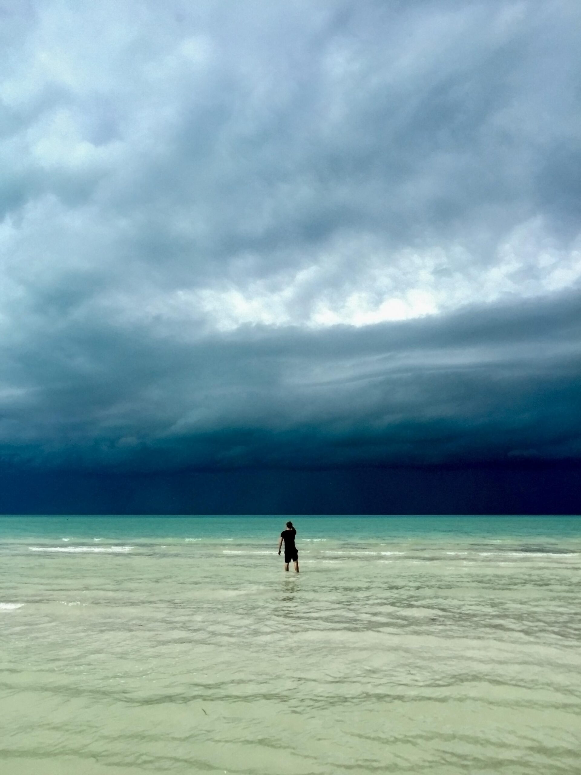 A person in black attire stands on a serene beach under dramatic, cloudy skies in Mexico.
